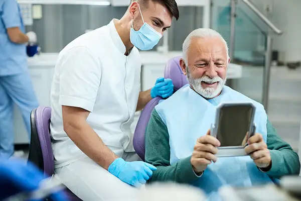 Smiling senior patient with dental implants looking at himself in a hand mirror
